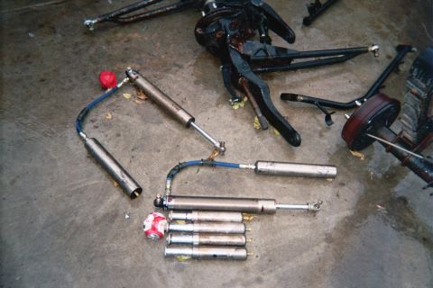 1990 Ford Bronco - tie-rod steering linkage and several shock absorbers with blue hoses lie on a concrete garage floor near an axle assembly.