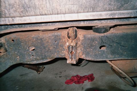 1990 Ford Bronco - Underside view of a rusty steel crossmember and frame rails of a vehicle, with a red rag on the floor below.