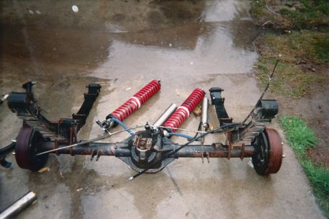 1990 Ford Bronco - solid rear axle with brake drums lies on wet pavement alongside red-spring coilover shocks and a driveshaft.