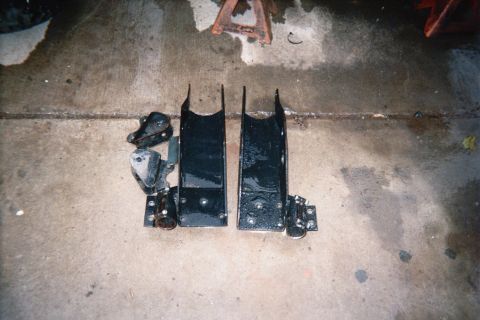 1990 Ford Bronco - Two freshly black-painted fabricated steel suspension brackets sit side by side on a concrete garage floor near jack stands.
