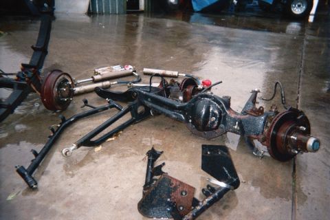 1990 Ford Bronco - solid axle assembly with black control arms, brake hubs and shocks rests on a wet garage floor during a suspension build.