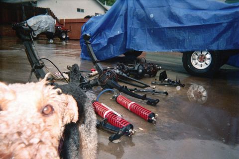 1990 Ford Bronco - Axles, a leaf spring and red coilover shocks lie on wet pavement beside a tarp-covered vehicle, with a dog's head in the foreground.