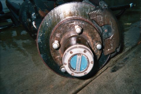1990 Ford Bronco - Close-up of a rusty front disc brake rotor with caliper and a blue manual locking hub mounted on the spindle.