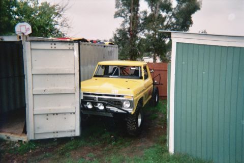 1990 Ford Bronco - yellow lifted Ford pickup with off-road lights and chrome wheels sits between a metal container and a green shed under trees.