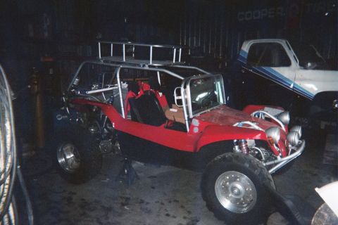 1990 Ford Bronco - red dune buggy with roll cage and coilover shocks sits on stands in a dark garage next to a blue pickup truck.