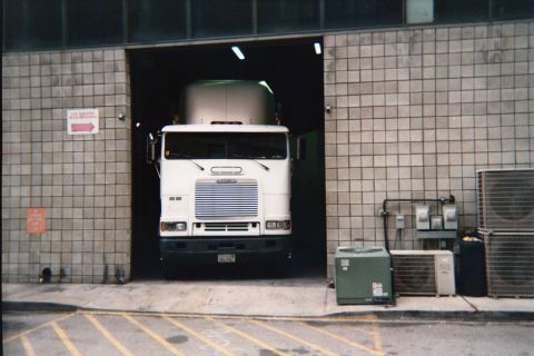1990 Ford Bronco - white cab-over semi-truck nosed into the open doorway of a tan tiled building beside outdoor HVAC condenser units.