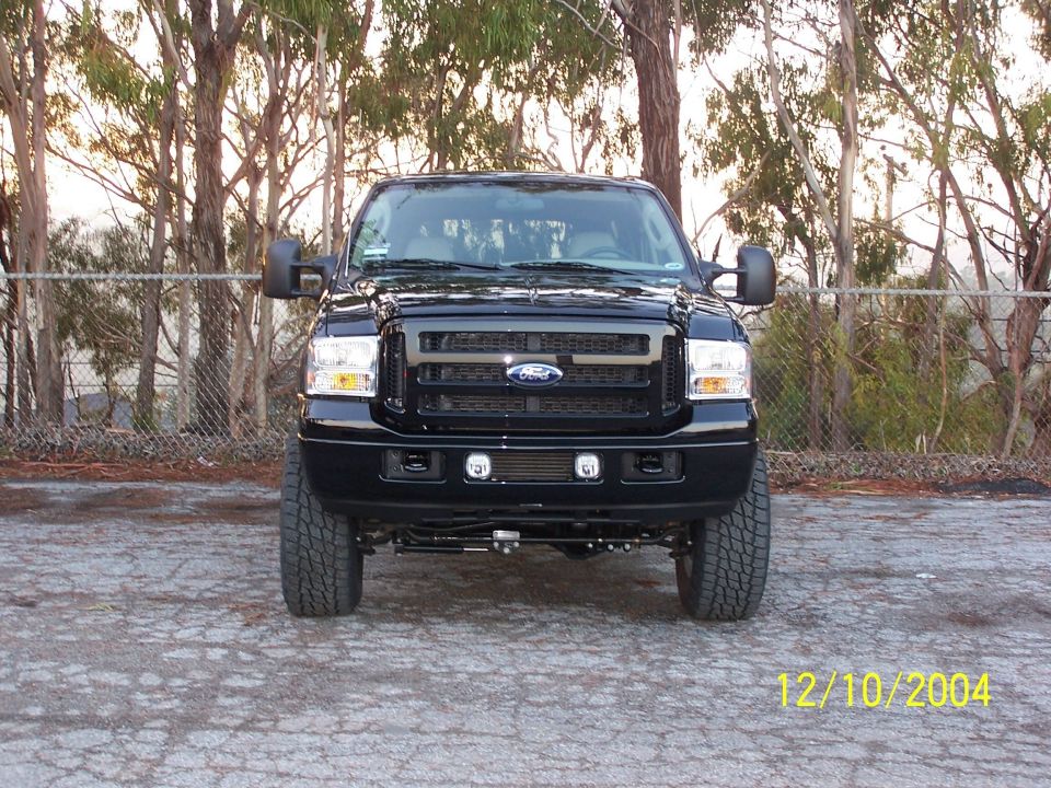 Front view of lifted black Ford Excursion with fog lights, parked by chain-link fence and trees