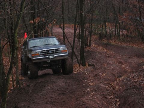 1995 Ford Bronco - A lifted 1995 Ford Bronco navigates a challenging muddy off-road course surrounded by trees.