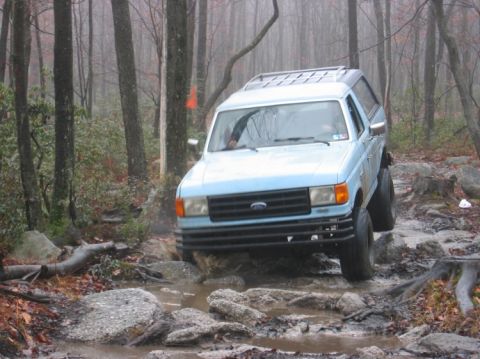 1995 Ford Bronco - This light blue SUV is shown off-roading through a challenging muddy and rocky path in a wooded area.