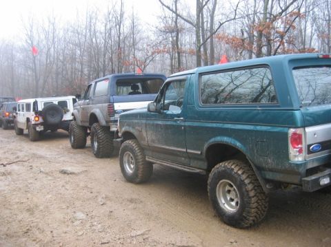 1995 Ford Bronco - Several SUVs, including a 1995 Ford Bronco, are lined up on a muddy trail in a wooded area.