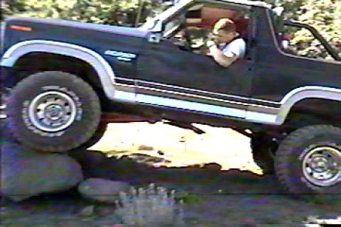 1984 Ford Bronco - Grainy side view of a dark blue lifted Bronco with its top removed and roll bar exposed, flexing over rocks at a water crossing with a driv…