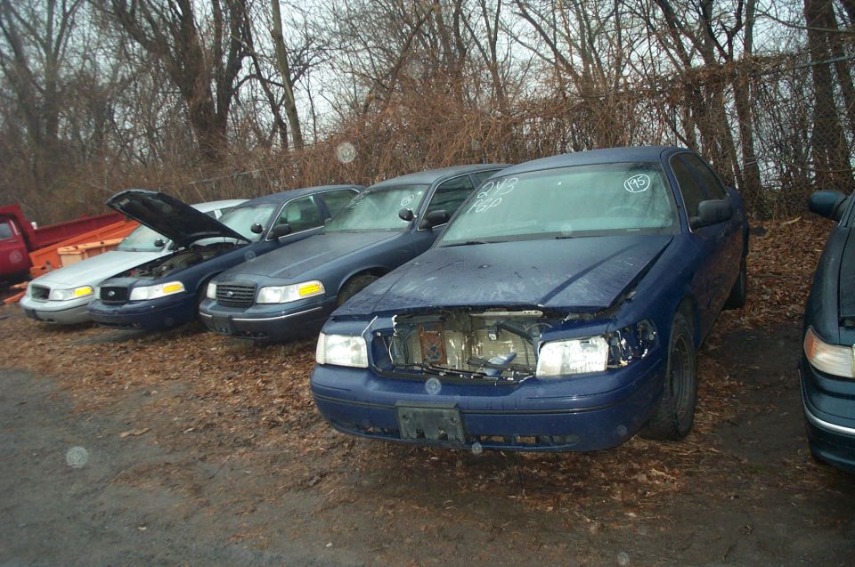 Blue Ford Crown Victoria with hood open, missing grille, parked in a lot with other vehicles