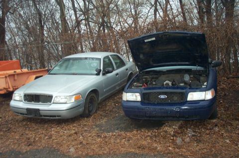 1991 Ford LTD Crown Victoria - Two Ford Crown Victorias are parked side-by-side in an outdoor lot, with the blue vehicle's hood open revealing the engine.