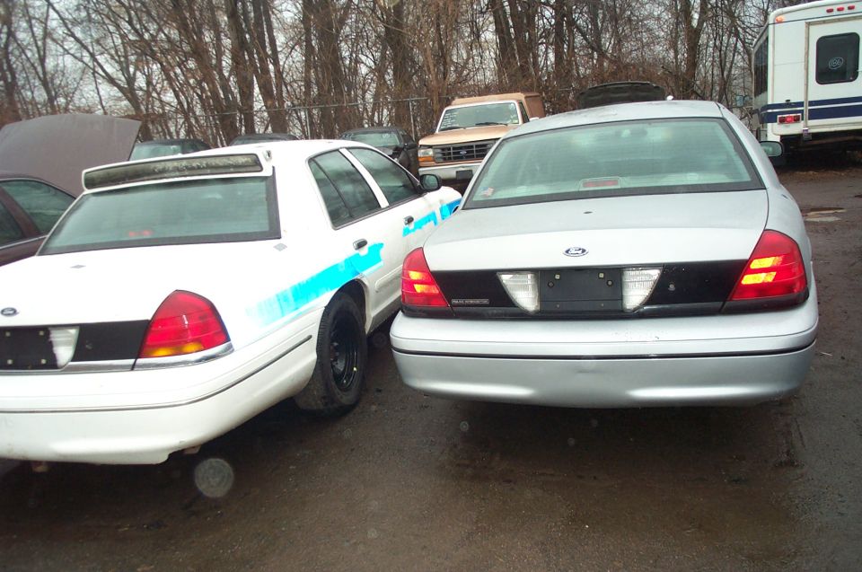 Two silver Ford Crown Victorias parked side-by-side, rear view. One has blue markings on its side