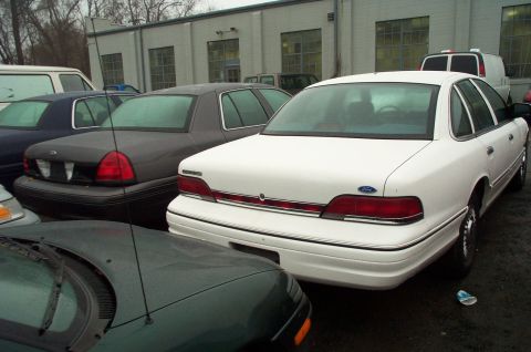 1991 Ford LTD Crown Victoria - Multiple sedans are parked in a lot near a building, including a white Ford Crown Victoria.