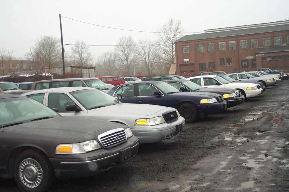 Several Ford Crown Victoria police cars parked in a lot, some dark blue, some silver. A brick building is in the background