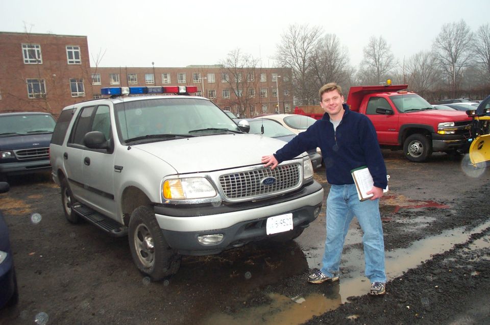 Man standing next to silver Ford Expedition with red and blue light bar on roof. Red dump truck with snow plow visible behind