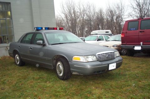 1991 Ford LTD Crown Victoria - Gray police car with light bar parked on grass. Other vehicles are visible in the background.