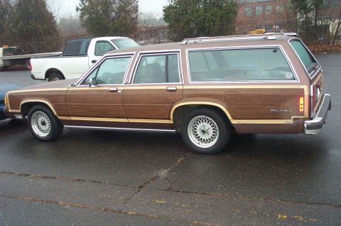 1991 Ford LTD Crown Victoria - A brown station wagon with wood paneling is parked on a wet asphalt lot next to a white pickup truck.