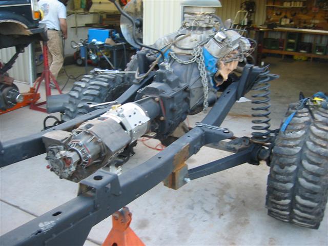 Vehicle chassis on jack stands with engine and transmission installed. A person stands in the background