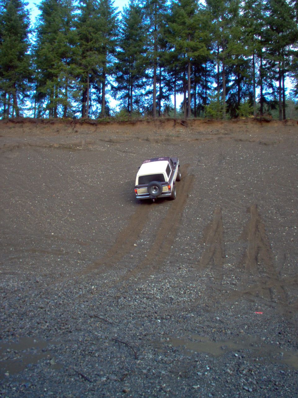 Rear view of two-tone burgundy and white Ford Bronco climbing a steep gravel hill with tire tracks, trees at top