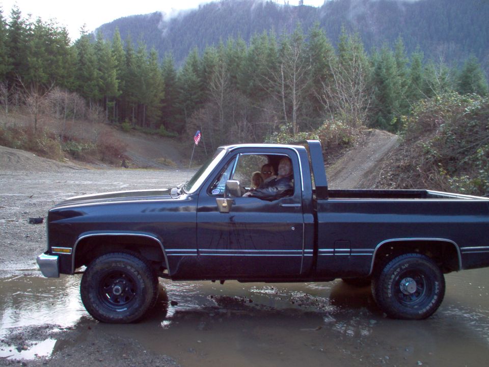 Dark blue short-bed pickup truck in profile on muddy gravel with forested hills behind