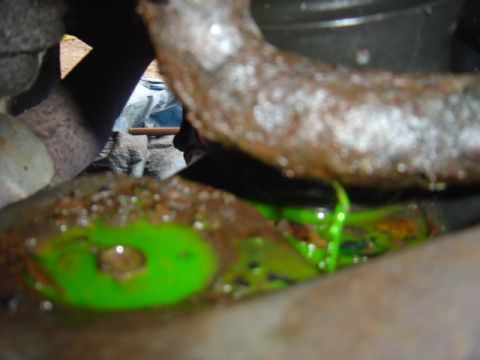 1998 Ford Taurus - Close-up under a vehicle showing bright green coolant dripping past a heavily rusted pipe and pooling on a rusty crossmember below, evidenc…