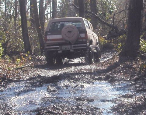 1988 Ford Bronco - A tan SUV navigates a muddy path through a wooded area, kicking up water and mud.