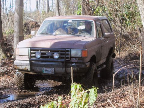 1988 Ford Bronco - A brown Ford Bronco navigates a muddy trail surrounded by trees.