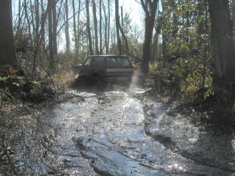 1988 Ford Bronco - A dark SUV navigates a muddy trail surrounded by trees, kicking up water and spray.