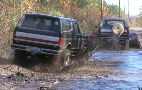 1988 Ford Bronco - Two Ford Broncos navigate a muddy trail, splashing water and dirt as they go.