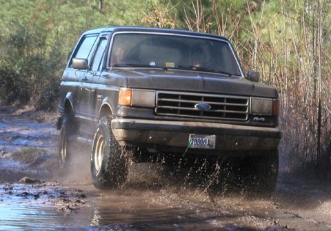 1988 Ford Bronco - A dark gray Ford Bronco navigates a muddy terrain, kicking up water and dirt as it drives forward.