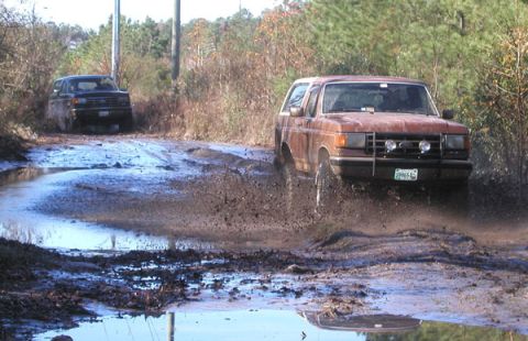 1988 Ford Bronco - Two SUVs navigate a muddy track, with the lead vehicle kicking up a large spray.
