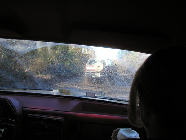 View from inside a car looking out the windshield at another SUV with a spare tire on the back, driving on a dirt road