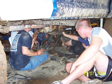 1979 Ford F-150 - Several individuals are gathered beneath a blue truck, appearing to perform maintenance or repairs on its undercarriage.