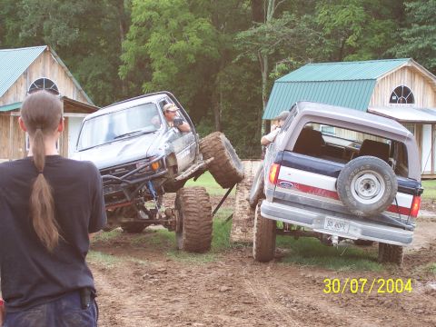 1979 Ford F-150 - Two heavily modified trucks are engaged in off-roading activities in a muddy area, with buildings in the background.