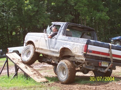 1979 Ford F-150 - A man drives a lifted, muddy Ford Bronco up a wooden ramp during an off-road event.