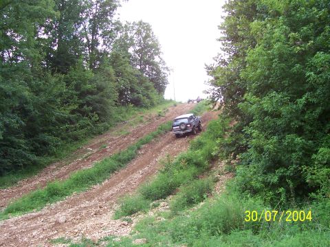 1979 Ford F-150 - A dark SUV navigates a steep, rutted dirt incline surrounded by trees, with other vehicles visible further up the hill.