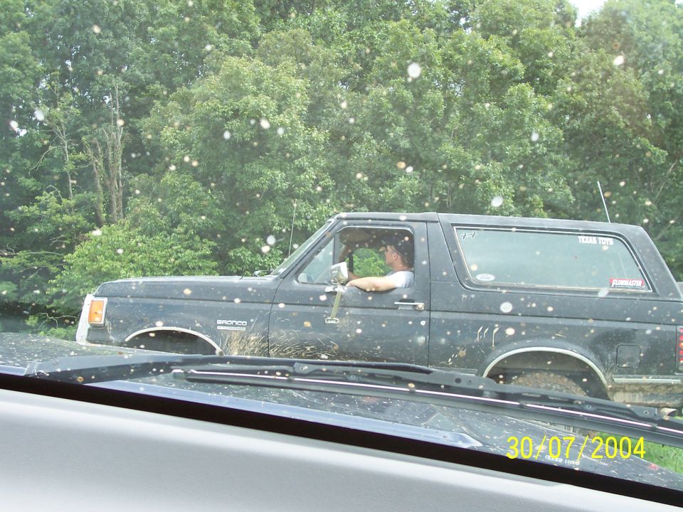 Black Ford Bronco driving off-road, driver visible. Another vehicle in foreground