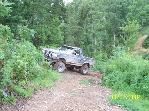1979 Ford F-150 - A lifted, muddy pickup truck navigates a dirt path through a wooded area on a summer day.