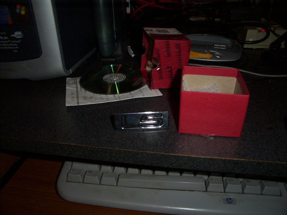 Chrome emblem with 'S' on a desk next to a CD and red box