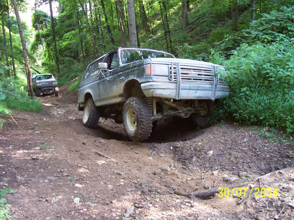 Muddy Ford Bronco climbing a dirt trail, another SUV behind it in the woods