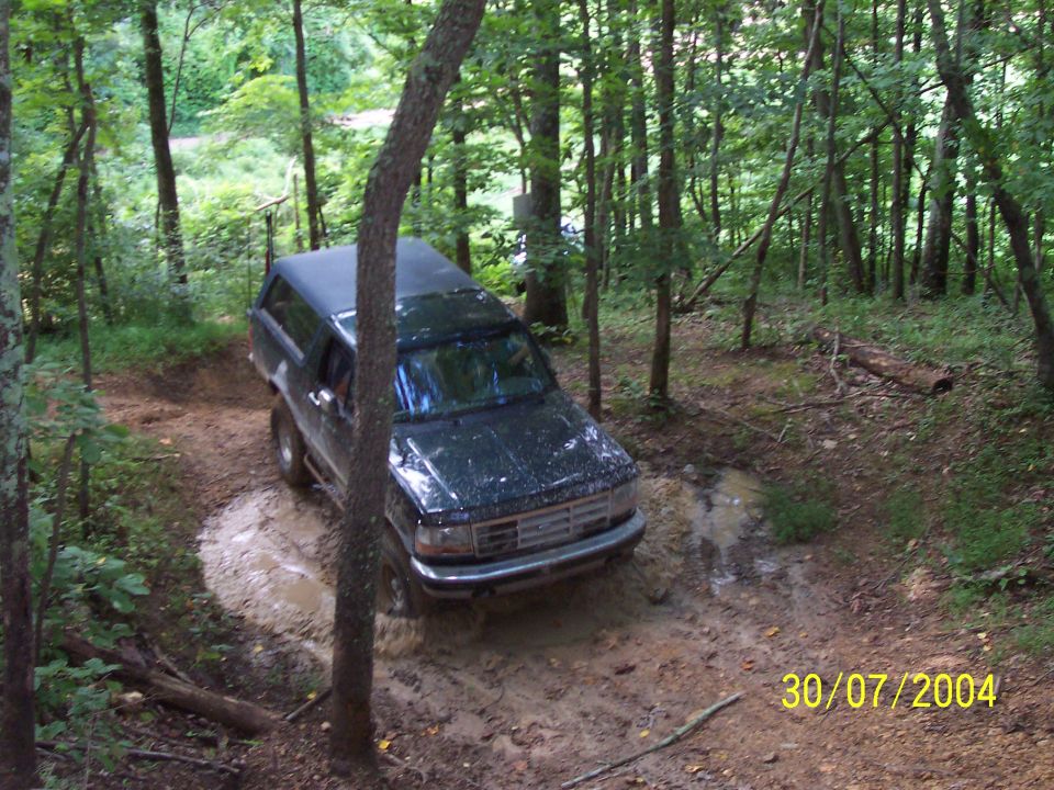 Dark green Ford Bronco navigating a muddy trail in a forest. Date stamp reads 30/07/2004