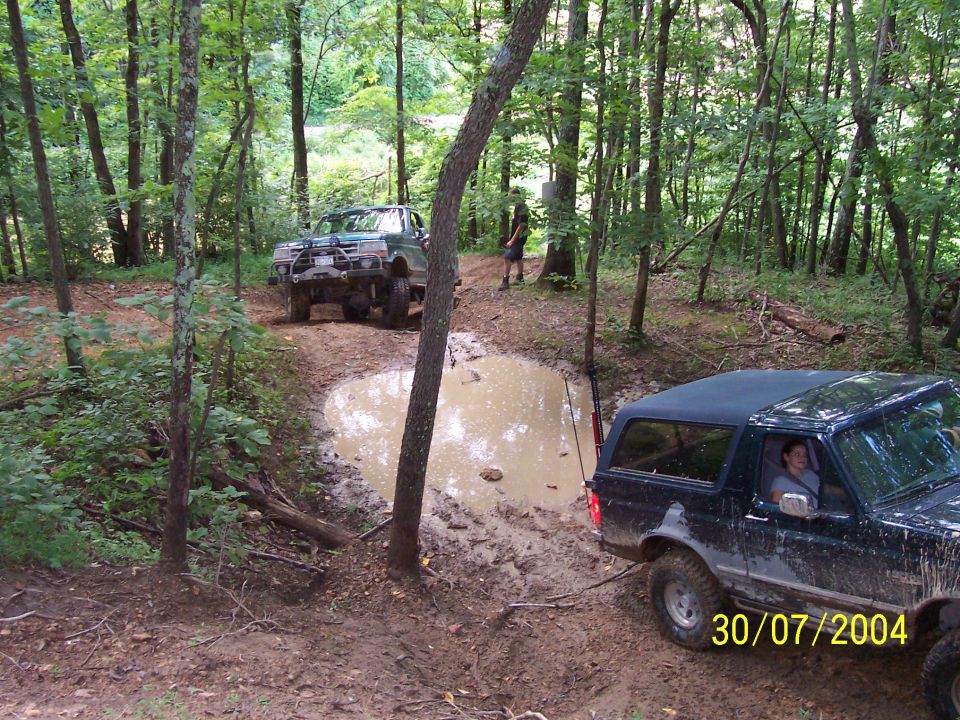 Two off-road trucks navigate a muddy path with a large puddle in a wooded area. A date stamp reads 30/07/2004