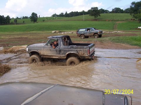 1979 Ford F-150 - Two off-road vehicles navigate a muddy terrain, with one Bronco deeply stuck in the water.