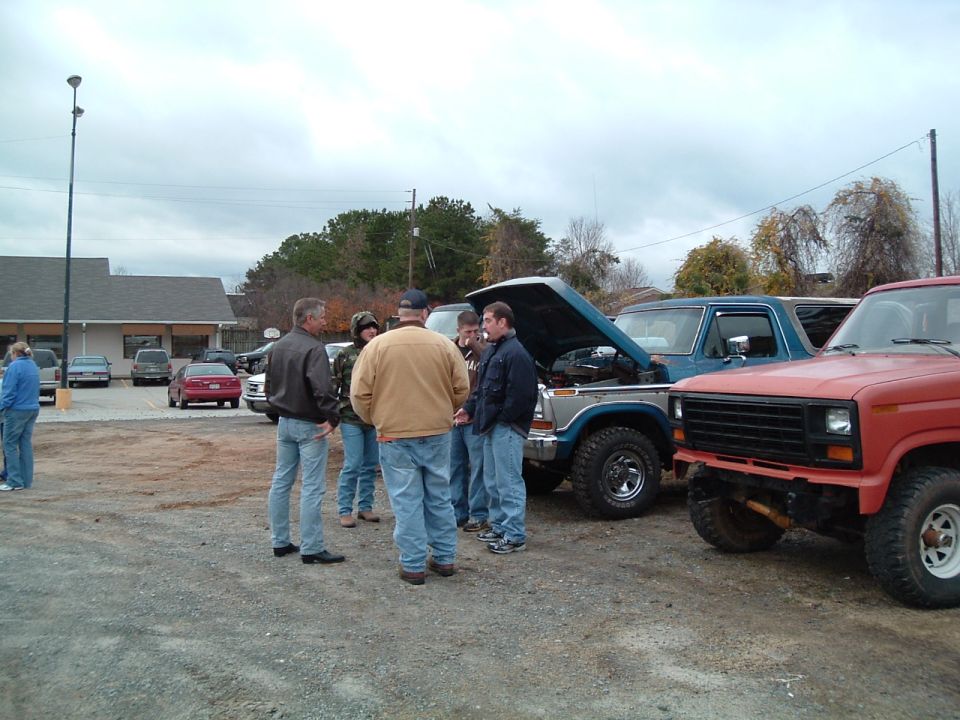 People gathered around a blue and silver Ford Bronco with hood up in a gravel lot, red Ford truck in foreground