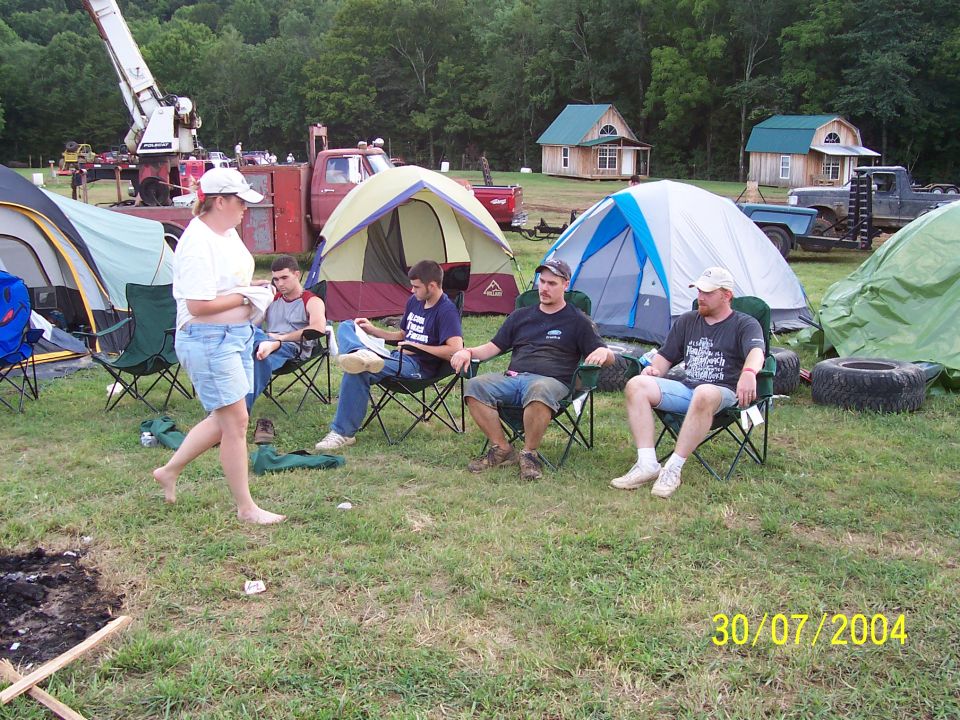 Campground scene with people sitting in chairs near tents, a red truck with a crane, and other vehicles in the background