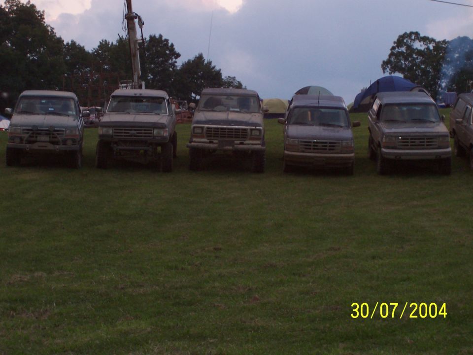 Five trucks parked in a grassy field with tents and trees in the background