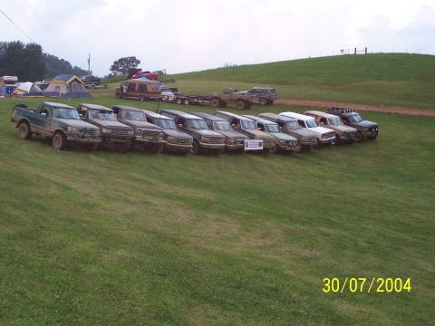 1979 Ford F-150 - Multiple pickup trucks and SUVs are parked in a line on a grassy slope, with camping tents visible in the background.