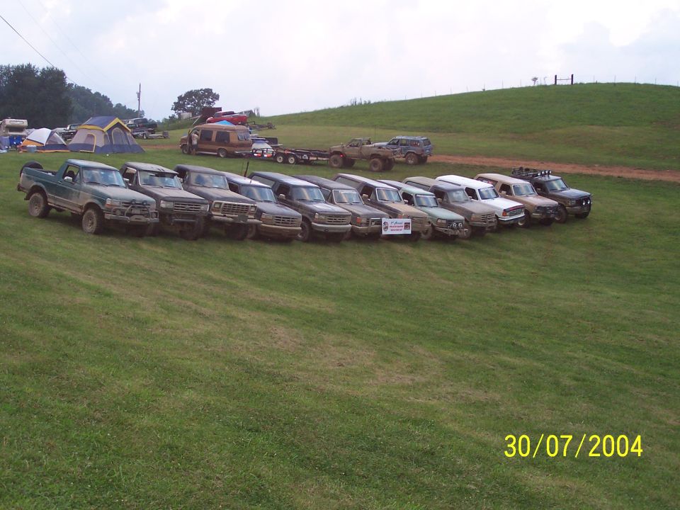 Line of pickup trucks and SUVs parked on a grassy hill, with tents and a van in the background. Date stamp reads 30/07/2004